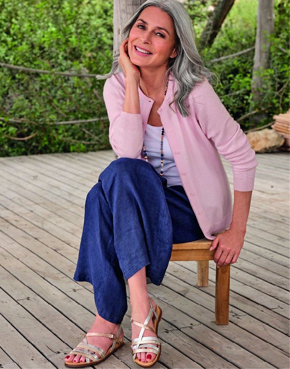A woman with grey hair, wearing a pink cardigan, smiling on a wooden deck.